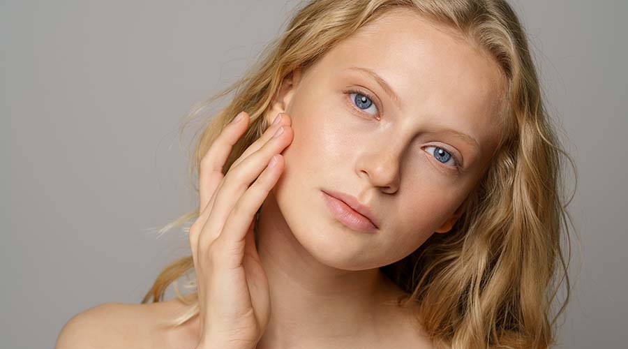 Belle femme blonde de face se touche le visage et sa peau sensible sur fond beige et gris